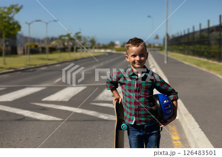 Smiling boy holding skateboard and helmet standing on school road, copy space 126483501