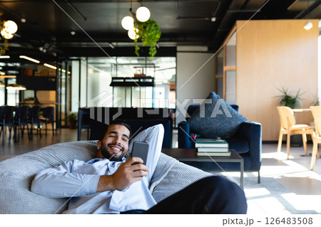 Relaxing in modern office, man smiling while using smartphone on bean bag 126483508
