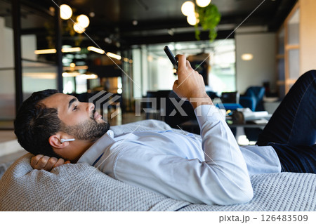 Relaxing in office lounge, man using smartphone and wearing wireless earbuds 126483509