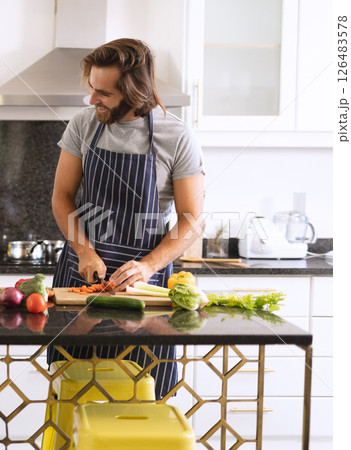 Man in kitchen chopping vegetables on counter, wearing apron, smiling while cooking 126483578