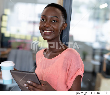 Smiling woman holding tablet and coffee cup in modern office environment 126483584