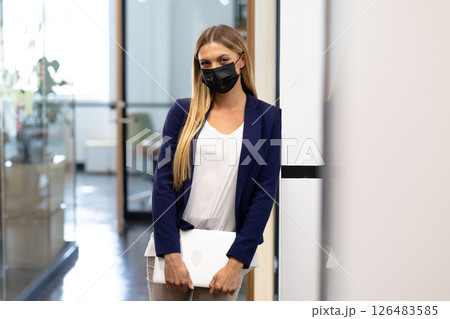 Professional woman in office holding laptop, wearing mask, looking confident 126483585