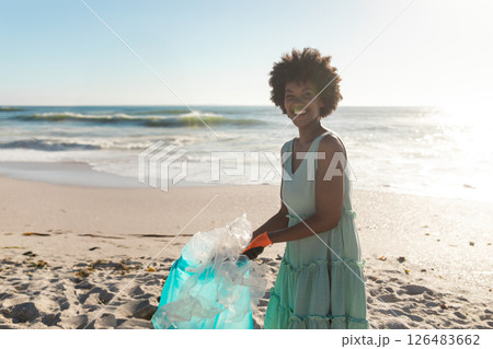 African American woman cleaning beach, collecting plastic waste, smiling in sunlight 126483662
