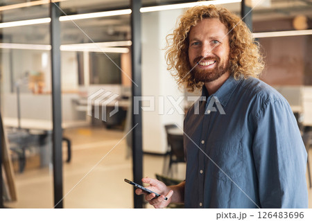 Smiling man with smartphone standing in modern office, looking confident, copy space 126483696