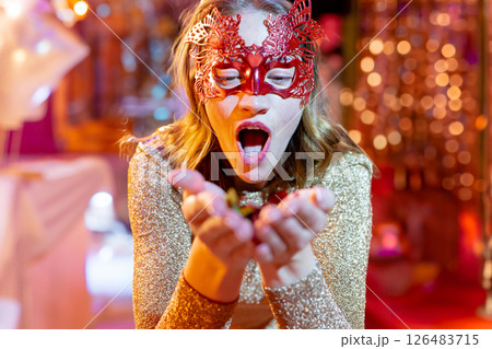 Woman in glittery dress wearing red mask, joyfully holding confetti at party Woman in glittery dress wearing red mask, joyfully holding confetti at party 126483715