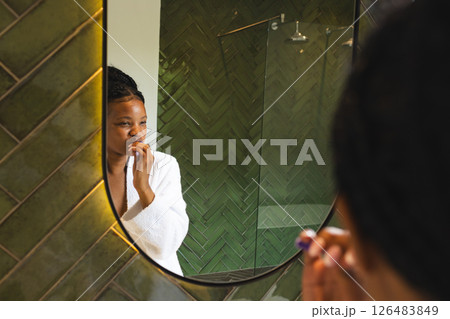 Woman in white robe brushing teeth in modern bathroom, reflecting in mirror 126483849