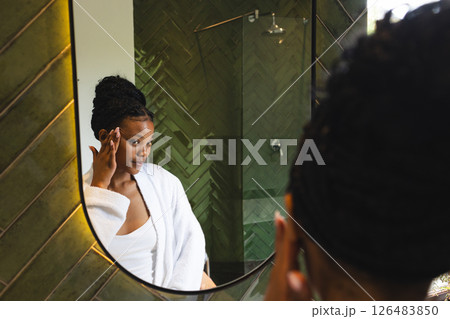 Woman in bathroom applying skincare, wearing white robe, looking in mirror 126483850