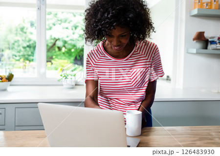 Smiling woman in striped shirt using laptop and holding coffee mug in kitchen Smiling woman in striped shirt using laptop and holding coffee mug in kitchen 126483930