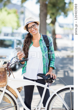 Smiling woman with smartphone standing beside bicycle on sunny city street, copy space 126483969