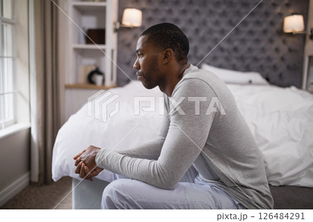 Contemplating life, African American man sitting on bed, gazing out window Contemplating life, African American man sitting on bed, gazing out window 126484291