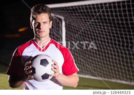 Soccer player holding ball confidently on field in front of goalpost at night, copy space 126484323