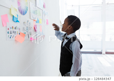 Young boy in office analyzing colorful charts on wall, wearing formal attire, copy space 126484347