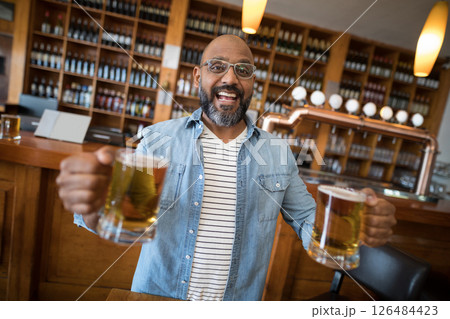 Smiling man holding beer mugs in lively bar with wooden shelves 126484423