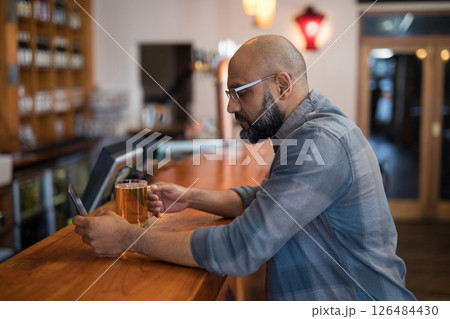 Man enjoying beer while browsing smartphone at cozy bar counter, copy space 126484430