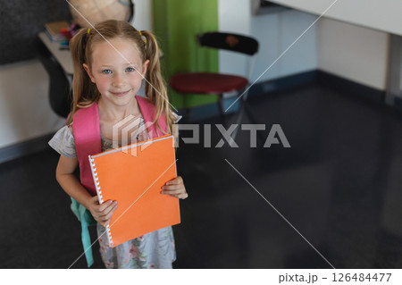 Smiling girl holding orange notebook in classroom, ready for school day, copy space 126484477