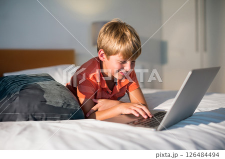 Young boy using laptop on bed at home, smiling and enjoying online activity 126484494