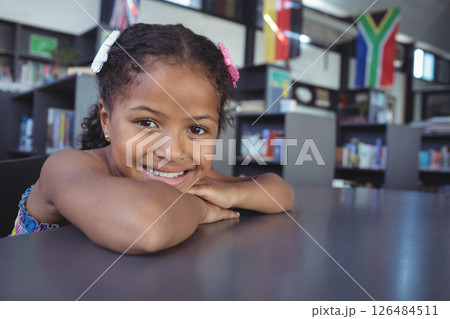 Smiling girl in school library resting on table, surrounded by books and flags, copy space 126484511