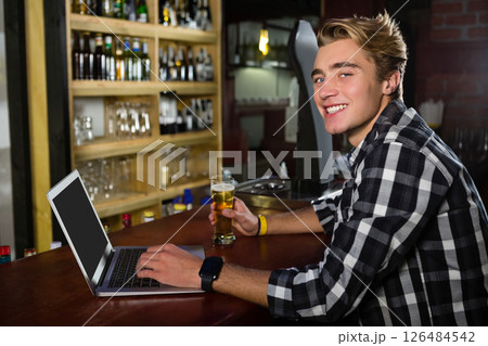Young man using laptop and enjoying beer at bar, smiling at camera, copy space Young man using laptop and enjoying beer at bar, smiling at camera, copy space 126484542
