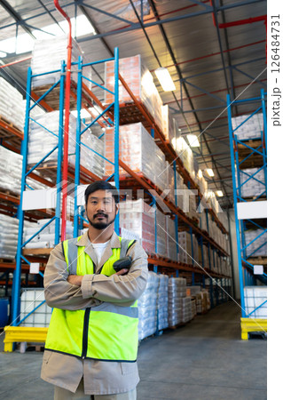 Warehouse worker in safety vest standing confidently in front of tall shelves, copy space 126484731