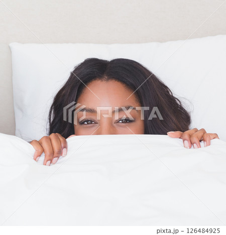 Smiling woman peeking from under blanket in cozy bedroom setting Smiling woman peeking from under blanket in cozy bedroom setting 126484925