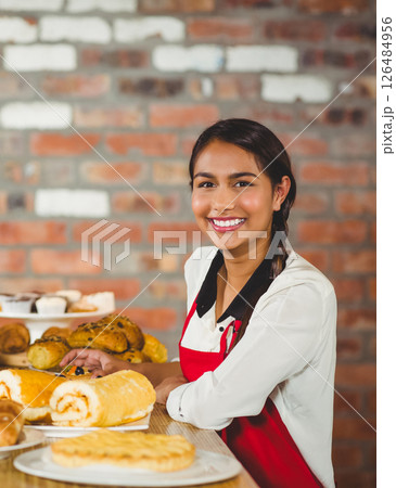 Smiling woman in red apron presenting fresh pastries at bakery counter 126484956