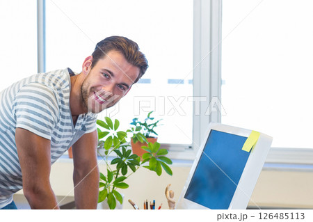 Smiling man leaning on desk in office, working with computer and plant Smiling man leaning on desk in office, working with computer and plant 126485113