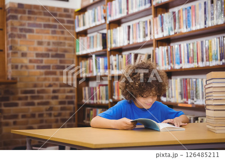 Curly-haired boy reading book in library, surrounded by shelves of books 126485211
