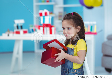 Smiling girl holding gift box in colorful playroom, feeling excited and curious 126485229