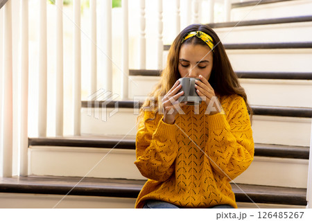 Woman in yellow sweater enjoying coffee on staircase at home, feeling relaxed, copy space 126485267