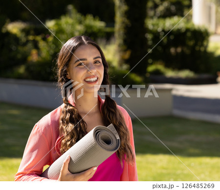Smiling woman holding yoga mat outdoors, ready for exercise in garden, copy space 126485269