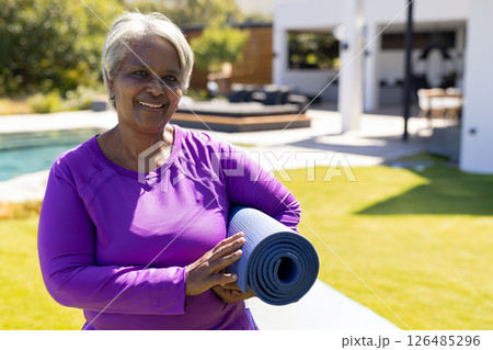 Senior woman holding yoga mat, smiling by poolside, enjoying outdoor relaxation, copy space 126485296