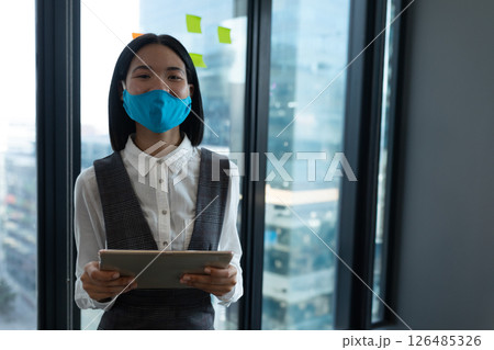 Businesswoman wearing mask holding tablet in modern office, looking confident, copy space Businesswoman wearing mask holding tablet in modern office, looking confident, copy space 126485326