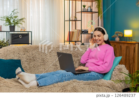 Young woman sitting on sofa wearing headphones listening music while working on laptop with feet up 126489237