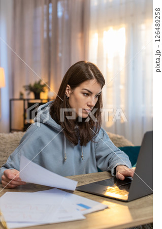 Focused woman freelancer reading documents and using laptop computer at table in living room at home 126489258
