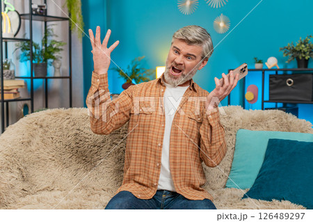 Mature man having phone argument on speaker, expressing stress and negative emotions on sofa at home 126489297