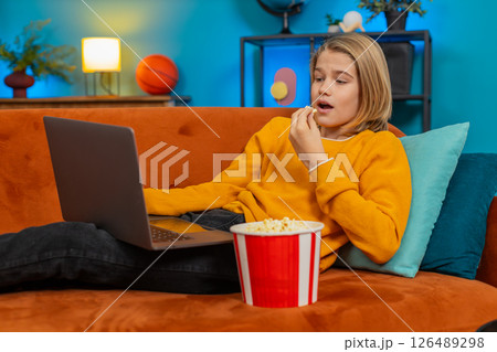 Teenager boy enjoying movie on laptop, eating popcorn, laughing at funny scene lying on sofa at home Teenager boy enjoying movie on laptop, eating popcorn, laughing at funny scene lying on sofa at home 126489298