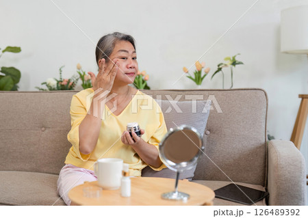 Beauty and Age Diversity. A senior woman applying skincare in a cozy living room setting. 126489392