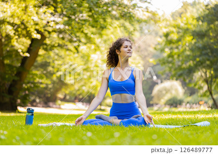 Woman sitting on mat in lotus position, relaxing practicing yoga meditation in park on summer day 126489777