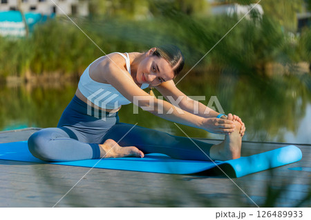 Fit Caucasian young woman in sportswear doing stretching exercising on yoga mat near pond in park 126489933