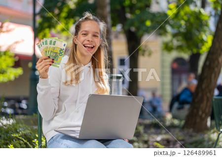 Happy rich woman using laptop winner victory holding cash money in euro banknotes sitting on chair 126489961