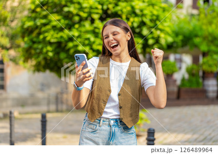 Happy Caucasian young woman using smartphone celebrating win good message news on city town street Happy Caucasian young woman using smartphone celebrating win good message news on city town street 126489964