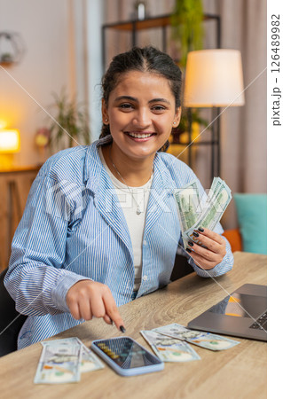 Happy rich Indian freelancer counting money cash using smartphone calculator app at home office desk Happy rich Indian freelancer counting money cash using smartphone calculator app at home office desk 126489982