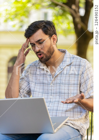 Displeased Indian man using laptop working, loses becoming surprised sudden lottery results outdoors Displeased Indian man using laptop working, loses becoming surprised sudden lottery results outdoors 126490152