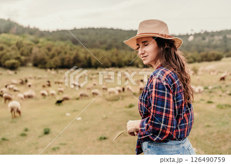 A woman wearing a straw hat stands in a field of sheep 126490759