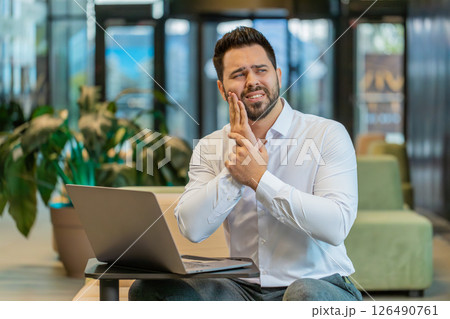 Caucasian businessman in office lobby working on laptop and feeling toothache in modern office lobby 126490761