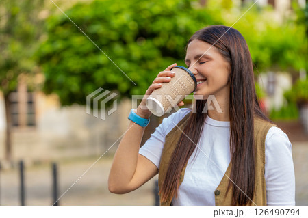 Happy Caucasian young pretty woman enjoying morning coffee hot drink and walking on city street Happy Caucasian young pretty woman enjoying morning coffee hot drink and walking on city street 126490794