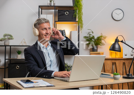 Happy young businessman in suit and wireless headphones enjoy music using laptop at home office desk Happy young businessman in suit and wireless headphones enjoy music using laptop at home office desk 126490820
