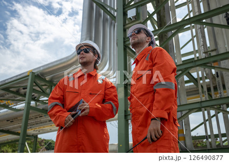 Two workers supervisor dressed in orange safety suits and white helmets observing pipeline project at construction site. engineer at industrial tube oil petrochemical. Two workers supervisor dressed in orange safety suits and white helmets observing pipeline project at construction site. engineer at industrial tube oil petrochemical. 126490877