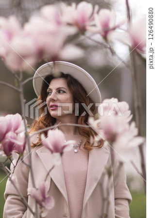 Woman magnolia flowers, surrounded by blossoming trees., hair down, white hat, wearing a light coat. Captured during spring, showcasing natural beauty and seasonal change. 126490880