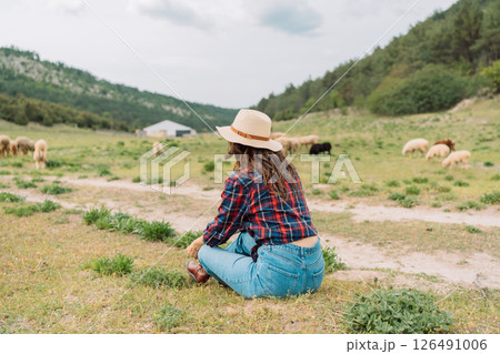 A woman in a plaid shirt and hat sits in a field with a herd of sheep 126491006